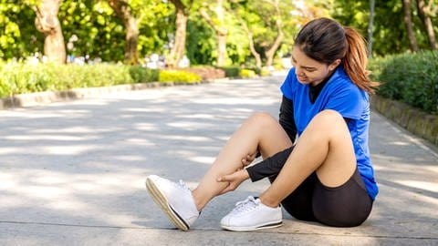 Eine Sportlerin in Joggingschuhe sitzt mit Schmerzen in der Wade auf dem Boden. Sie hat einen Muskelkrampf.