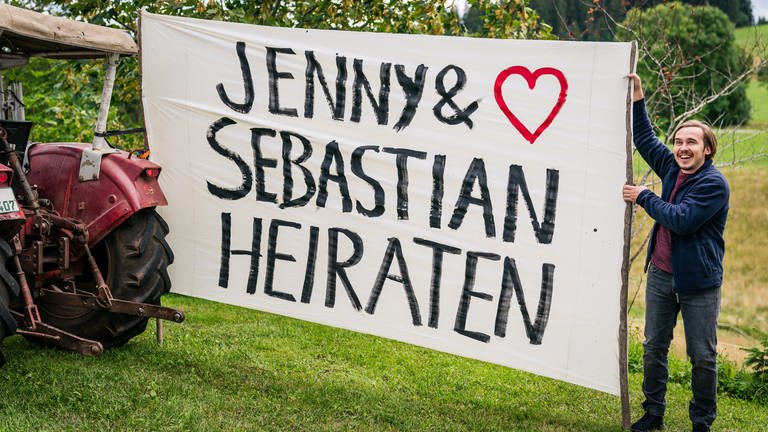 Sebastian steht neben einem riesigen Plakat mit der Aufschrift "Jenny und Sebastian heiraten"