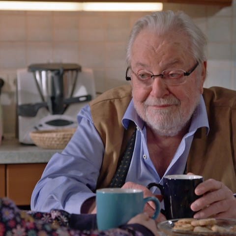 Hermann, der eine Kaffeetasse in der Hand hält, sitzt seiner Urenkelin, die man nur im Anschnitt von hinten sieht, am Küchentisch gegenüber.
