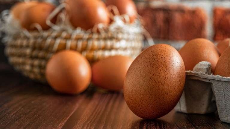 Im Vordergrund lehnt ein braunes Ei an einem vollen Eierkarton. Im Hintergrund sind weitere Eier in einem Strohkorb zu sehen.