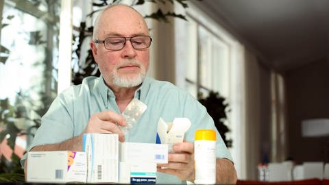 Ein älterer Mann sitzt an einem Tisch und studiert die Verpackung eines Medikamentes.