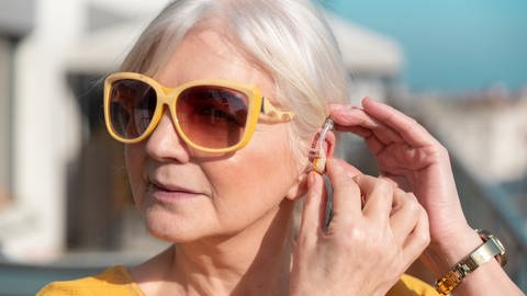 Eine ältere Dame klemmt sich ein Hörgerät hinters Ohr.