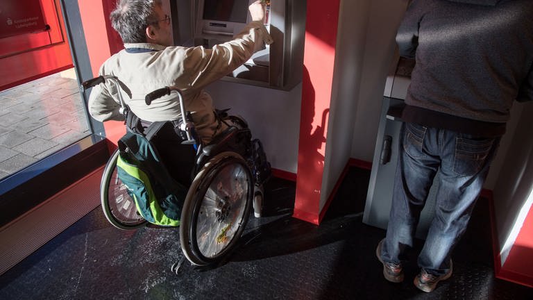 Mann im Rollstuhl hebt Geld an Geldautomat in Bankfiliale ab. Vorsicht bei Shoulder Surfing: Betrüger spähen dabei die PIN aus.
