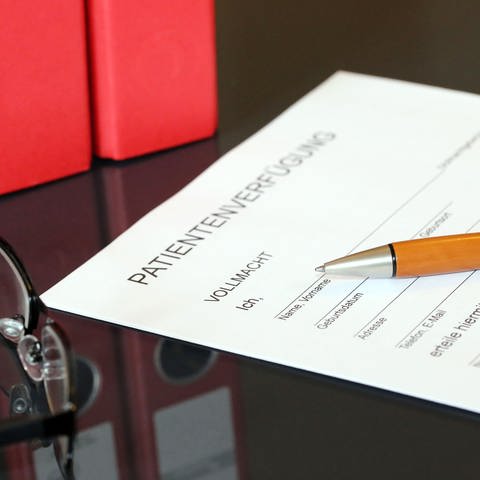 Ein Blatt Papier mit der Überschrift Patientenverfügung liegt auf einem Tisch. Auf dem Dokument liegt ein Kugelschreiber. Daneben auf dem Tisch liegt eine Brille. Dahinter stehen zwei Ordner.
