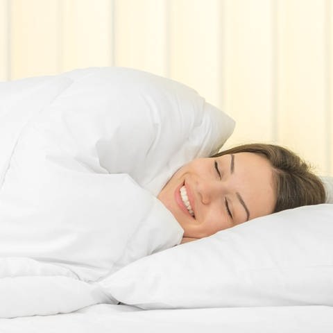 Eine Frau liegt im Bett. Ihr Kopf liegt auf einem Kissen. Ihr gesamter Körper ist von der Bettdecke umhüllt.