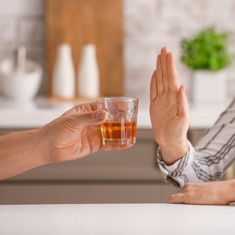 Zwei Menschen sitzen in einer Küche. Die linke Person hält in einer Hand ein Glas mit Alkohol und möchte das Getränk der anderen Person geben. Die Person auf der rechten Seite hält als "Stop-Signal" die Hand. Sie verweigert den Drink.