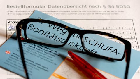 Auf dem Bestellformular für die Datenkopie der Schufa liegt eine Brille, ein roter Kugelschrieber und ein blauer Flyer mit der Aufschrift "Ihr Weg zur Schufa-Bonitätsauskunft".