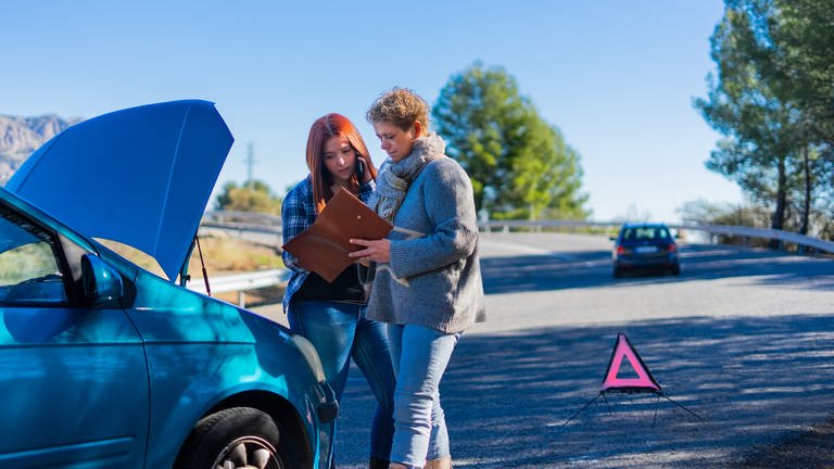 Zwei Frauen stehen vor der offenen Motorhaube eines Autos. Was tun, wenn man im Ausland an einem Autounfall beteiligt ist? Wer zahlt den Schaden? Und muss man Strafzettel aus dem Ausland bezahlen?