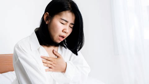 Asiatische Frau mit Panikstörung im Bett, die sich mit der Hand die Brust hält und kaum atmen kann