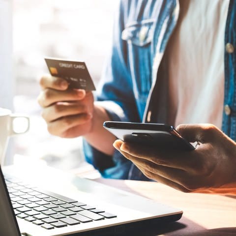 Person hält eine Bankkarte und ein Smartphone in der Hand und sitzt vor einem Laptop. Phishing: Wenn der Online-Shop Daten abgreift (Foto: Adobe Stock, Thapana_Studio)