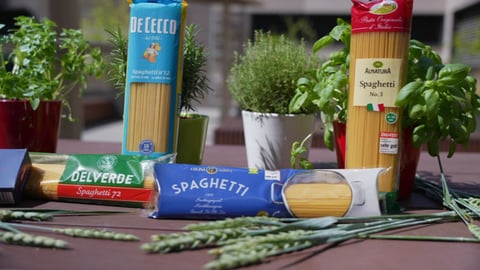 Spaghetti-Packungen der Marken De Cecco, Delverde, Alnatura und Aldi liegen zwischen Basilikumpflanzen auf einem Tisch.