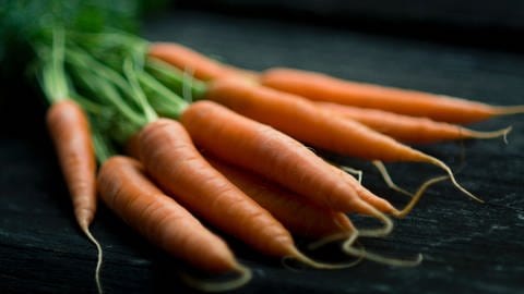 Zu sehen ist ein Bund Karotten auf einem Tisch. Karotten enthalten Beta-Carotin. Das ist eine Vorstufe von Vitamin A. Vitamin A benötigen wir für unseren normalen Sehvorgang.  In ihnen stecken jede Menge wertvoller Nährstoffe. Insbesondere die sogenannten Carotinoide, vor allem Beta-Carotin. Die natürlichen Farbstoffe geben der Karotte ihre Farbe und sind eine wichtige Quelle für Vitamin A.
