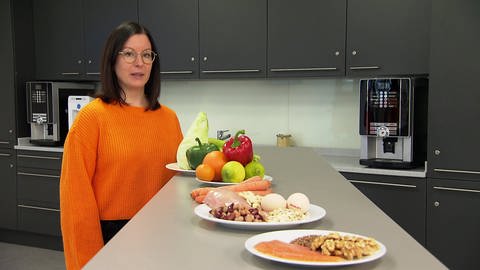 Christina Esser steht in einer Küche an einem Tisch auf dem drei Teller mit Gemüse, Obst Eiern, Hülsenfrüchten und Fisch sowie Fleisch stehen.