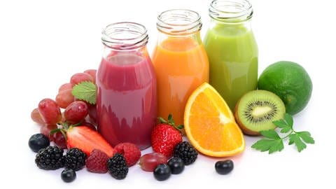 Drei Glasflaschen sind jeweils mit rotem, orangefarbenem und grünem Saft gefüllt. Um die Flaschen herum liegen verschiedene Früchte wie Erdbeeren, Orangen, Trauben, Beeren und Kiwi.