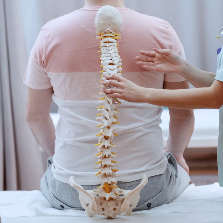 Gezeigt wird ein Patient, der mit dem Rücken zum Betrachtenden auf einer Liege sitzt. Eine Ärztin hält ein Skelettmodell einer Wirbelsäule an seinen Rücken.