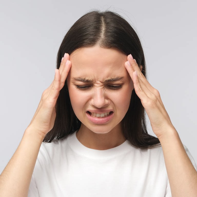 Junge Frau mit braunen glatten Haaren verzieht das Gesicht und hält sich die Schläfen. Was hilft bei Migräne? Neue Behandlungsmöglichkeiten, Tipps und Ursachen