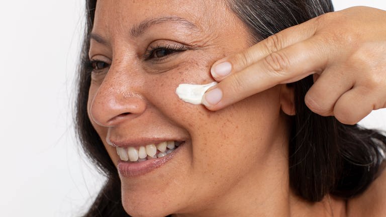 Frau mittleren Alters schmiert sich Creme ins Gesicht und lacht. Wie viel Hautpflege ist sinnvoll? Braucht man unbedingt eine Gesichtscreme?