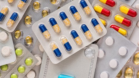 Es stapeln sich verschiedene Medikamentenverpackungen, in denen bunte Tabletten und Kapseln zu sehen sind. Für die Herstellung wird oft Gelatine aus Schlachtabfällen verwendet.