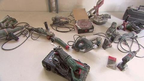 Mehrere Werkzeuge liegen auf einer Baustelle. Schleifmachine, Akkuschrauber, Schlagbohrer und weitere Geräte liegen auf dem Boden.