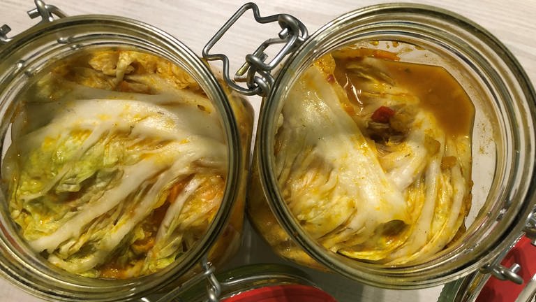 Blick von oben in zwei Drahtbügelgläser, deren Inhalt (Kimchi) mit einem Kohlblatt fest abgedeckt ist zum Fermentieren