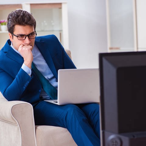 Ein Mann im Anzug mit einem Laptop auf dem Schoss schaut gebannt auf einen Fernseher (Foto: Colourbox)