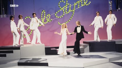 Auf einer weiß-schwarzen Bühne stehen im Zentrum Jule Styne und eine Frau. Von der Seitenbühne sind sie umrahmt von 5 Tänzern. (Foto: SWR, Annemie Huck)
