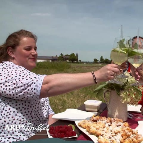 Drei Frauen stoßen mit Holunderblütensirup-Drinks an, auf dem Tisch: Hollerküchle