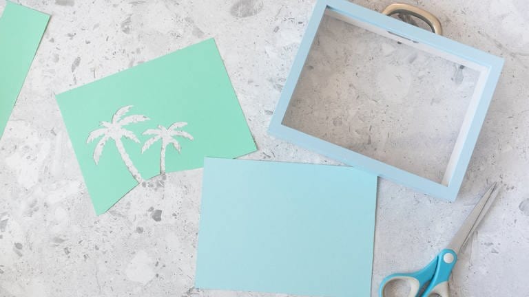 Aus einer grünen oder blauen Pappe die Silhouette von Palmen ausschneiden. De Pappe so zuschneiden, dass man sie vorne am Bilderrahmen einlegen kann. (Foto: Lisa Tihanyi)