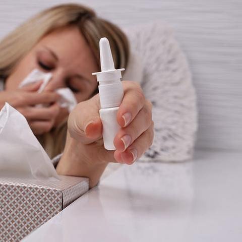 Eine Frau hält sich mit geschlossenen Augen ein Taschentuch vor Nase und Mund und greift nach einem Nasenspray (Foto: Getty Images, Thinkstock -)