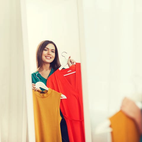 junge Frau steht mit Kleidungsstücken vor einem Spiegel