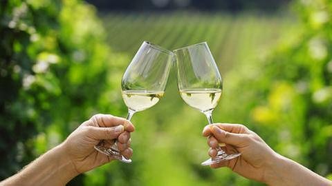Zei Personen prosten sich mit einem Glas Weißwein zu. Im Hintergrund sieht man unscharf Weinhänge. (Foto: Getty Images, Thinkstock -)