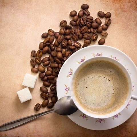 Eine Tasse Kaffee, daneben ein Löffel, Zucker und Kaffebohnen