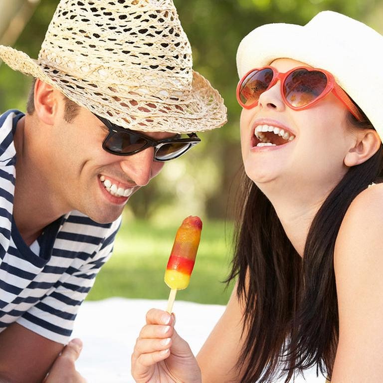 Junges Paar mit Sonnenhütten auf dem Kopf lacht und isst Eis