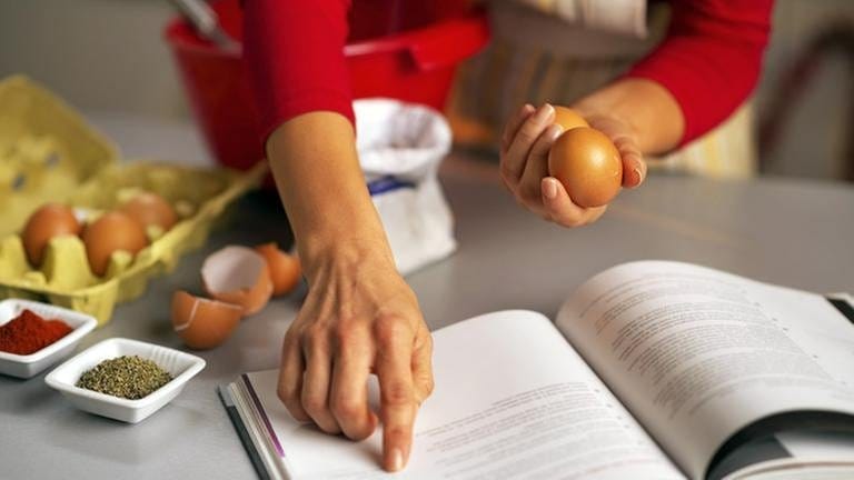 Eine Frau hat ein Kochbuch vor sich auf der Arbeitsplatte aufgeschlagen, diverse Zutaten stehen zur Verarbeitung bereit. (Foto: Getty Images, Thinkstock -)