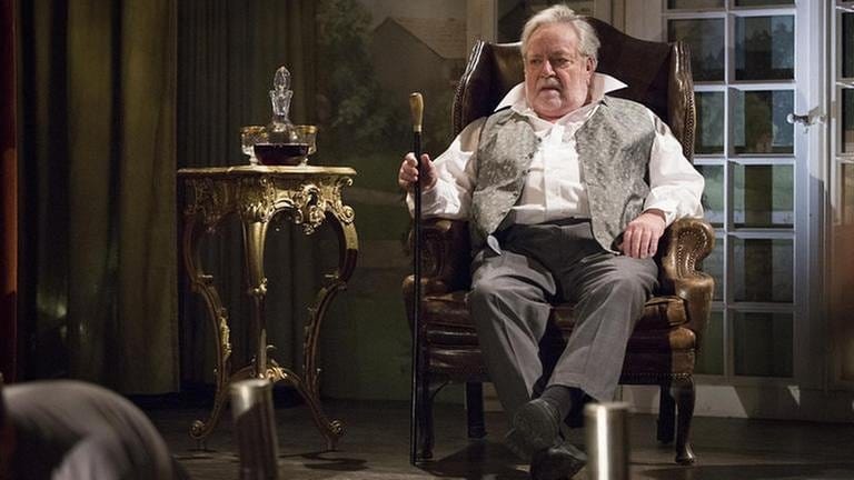 Hermann in einem großen Sessel auf der Theaterbühne