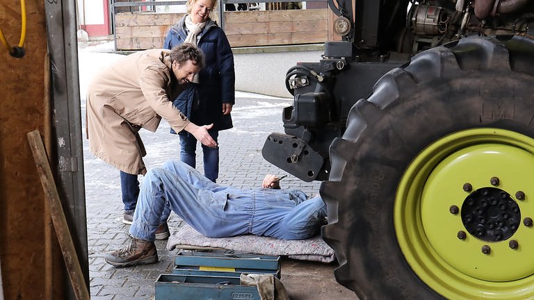 Karl liegt unter seinem Traktor, davor stehen Bea und ein Feriengast, der Karl begrüßen möchte (Foto: SWR, Johannes Krieg)