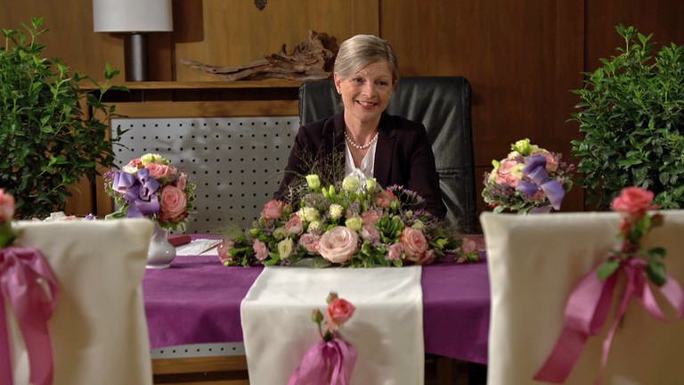 Frau Heilert sitzt an Bernhards Schreibtisch, der für eine standesamtliche Trauung geschmückt ist. (Foto: SWR)