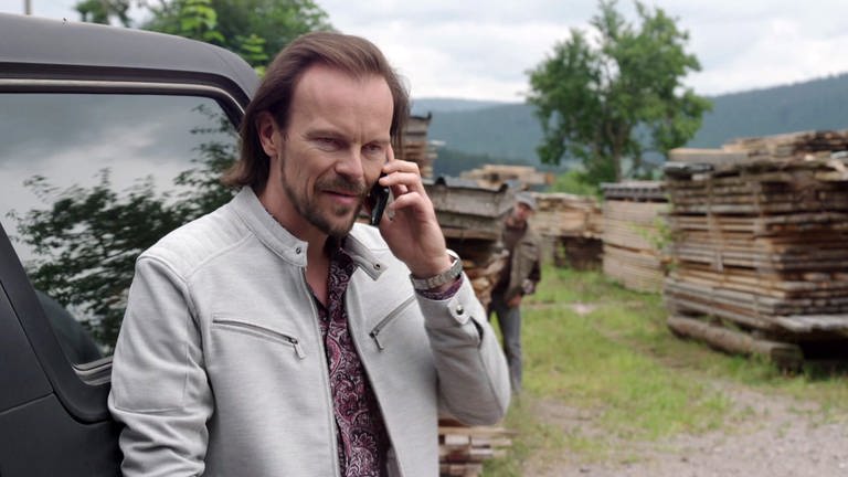 Constantin telefoniert, hinter einem Holzstapel steht Niki, der das Gespräch belauscht.  (Foto: SWR)