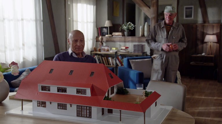Franz sitzt vor dem Modell seines geplanten Neubaus, Heinz steht in Stallkluft hinter ihm (Foto: SWR)