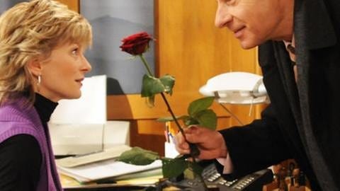 Franz überreicht Frau Heilert, die hinter ihrem Schreibtisch sitzt, eine rote Rose (Foto: SWR, SWR -)