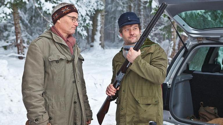 Karl und Riedle am verschneiten Waldrand, Riedle hält ein Gewehr hoch