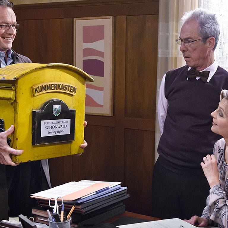 Bernhard bringt einen großen gelben Briefkasten ins Bürgermeisteramt, Frau Heilert und Herr Weiss staunen (Foto: SWR, SWR/Georg Zengerling -)