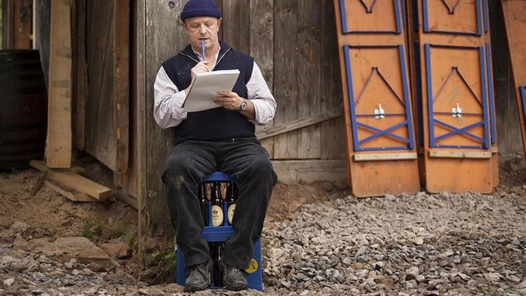 Toni sitzt mit Block und Bleistift auf einer Bierkiste vor der renovierten Mühle (Foto: SWR, SWR/Martin Furch -)
