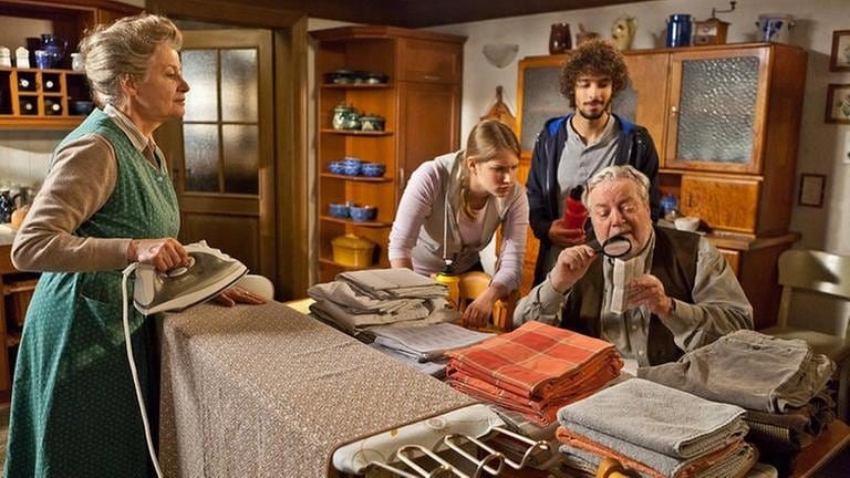Johanna bügelt in der Küche, Hermann sitzt am Küchentisch und schaut etwas mit einer Lupe an, Celine und Albert stehen hinter ihm (Foto: SWR, SWR/Peter Sebera -)