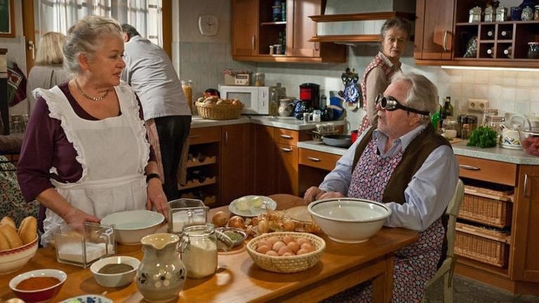 Leni werkelt am Küchentisch, Hermann sitzt mit Schutzbrille am Tisch und schneidet Zwiebeln, im Hintergrund steht Johanna am Herd