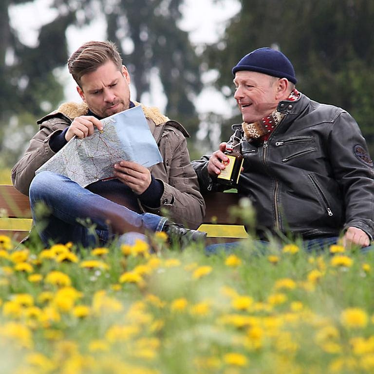 Andreas und Toni sitzen auf einer Bank, Toni hält ein Bier, Andreas studiert eine Landkarte, ringsum blüht der Löwenzahn (Foto: SWR, SWR/Johannes Krieg -)