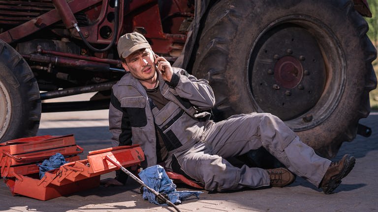 Sebastian telefoniert neben dem Traktor auf dem Boden sitzend (Foto: SWR, d:light / Christian Koch)