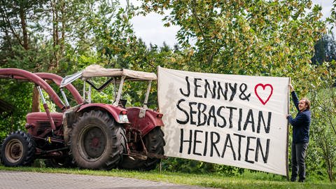 Sebastian hält ein riesiges Stück Stoff, das an einem Traktor befestigt ist. Darauf steht "Jenny und Sebastian heiraten"