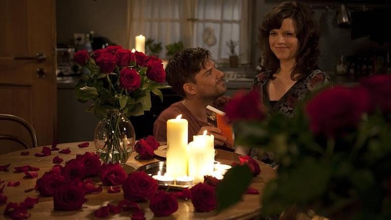 Andreas kniet vor Eva, die am Tisch sitzt, der mit Kerzen, einem Strauß roter Rosen und Rosenblättern geschmückt ist (Foto: SWR, SWR/Peter Sebera -)