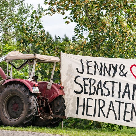 An einem roten Traktor befestigt und auf der anderen Seite von Sebastian gehalten ein großes Plakat auf dem steht: Jenny und Sebastian heiraten.